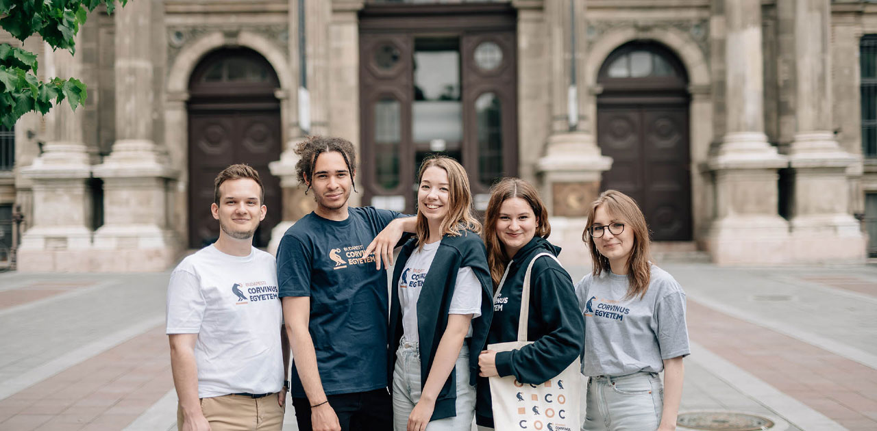 Corvinus University of Budapest önünde, üniversite logolu kıyafetler giyen beş öğrencinin birlikte poz verdiği grup fotoğrafı.