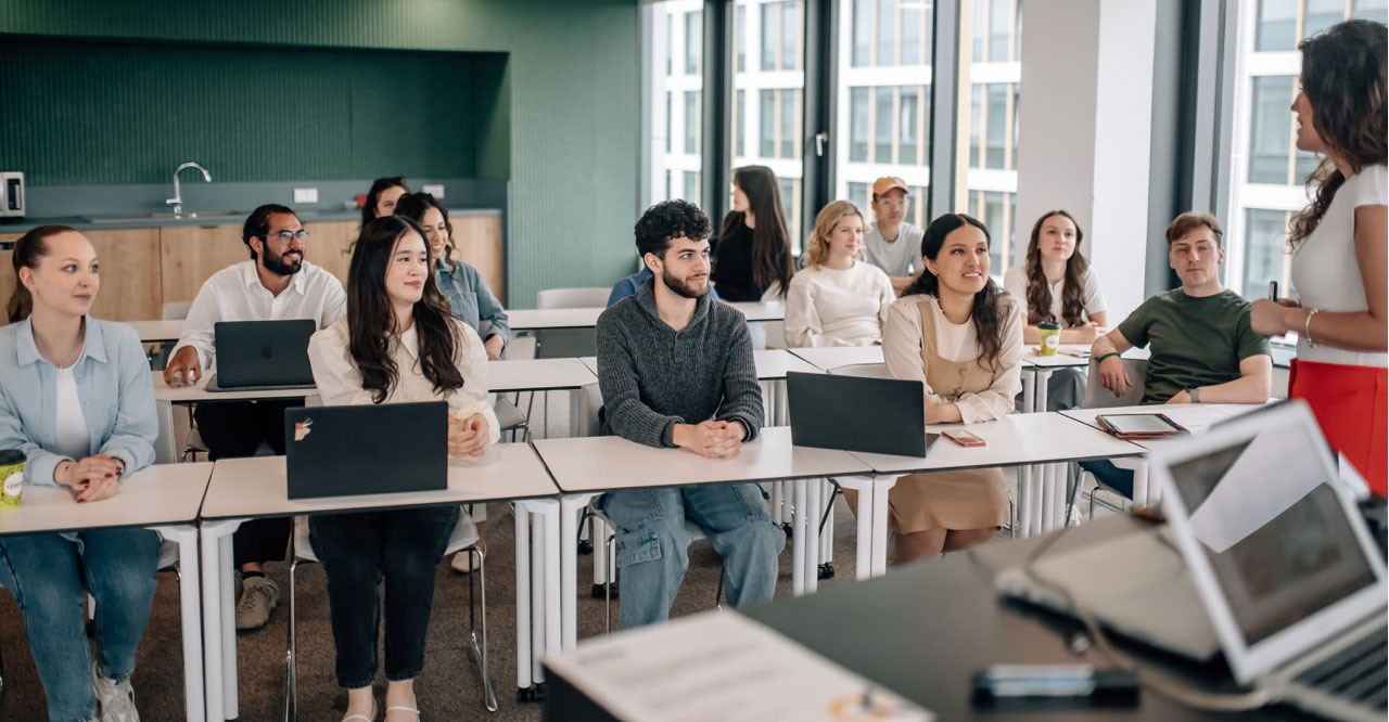 Modern bir sınıfta, dizüstü bilgisayarlarıyla derse odaklanmış çeşitli öğrenciler ve onlara hitap eden bir öğretim görevlisi