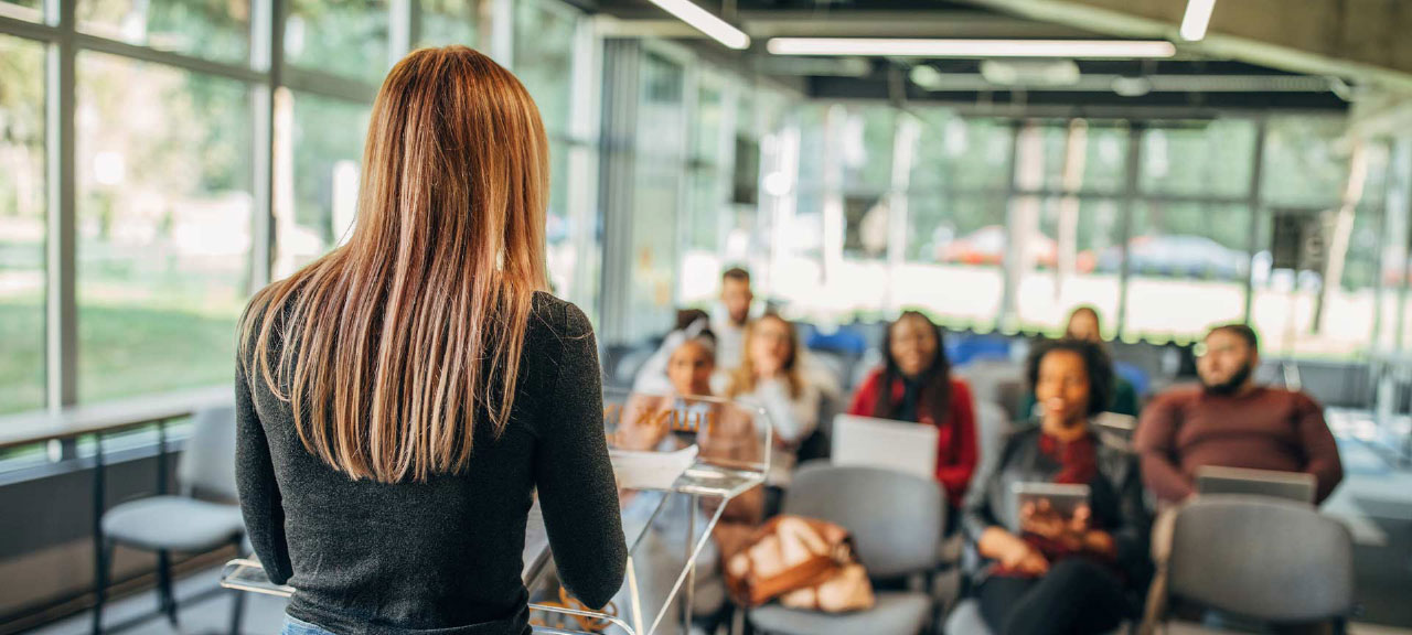 Cam duvarlarla çevrili modern bir alanda konuşmacıyı dinleyen bir grup öğrencinin yer aldığı kampüs etkinliği.