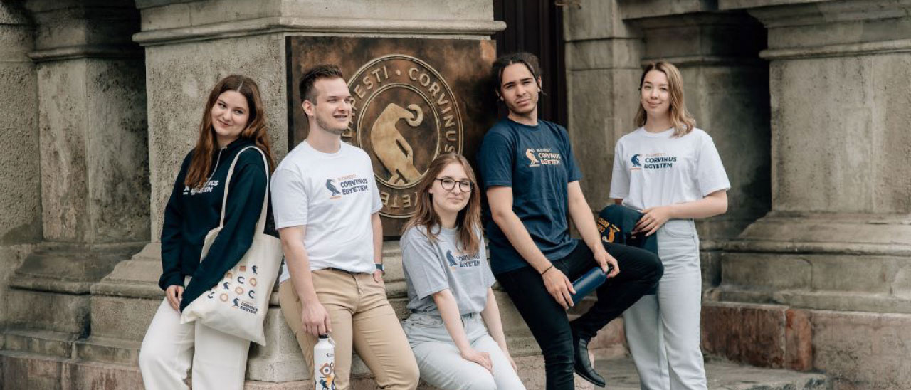 Corvinus University of Budapest ambleminin bulunduğu tarihi bina önünde üniversite logolu kıyafetler giyen beş öğrencinin birlikte poz verdiği grup fotoğrafı.