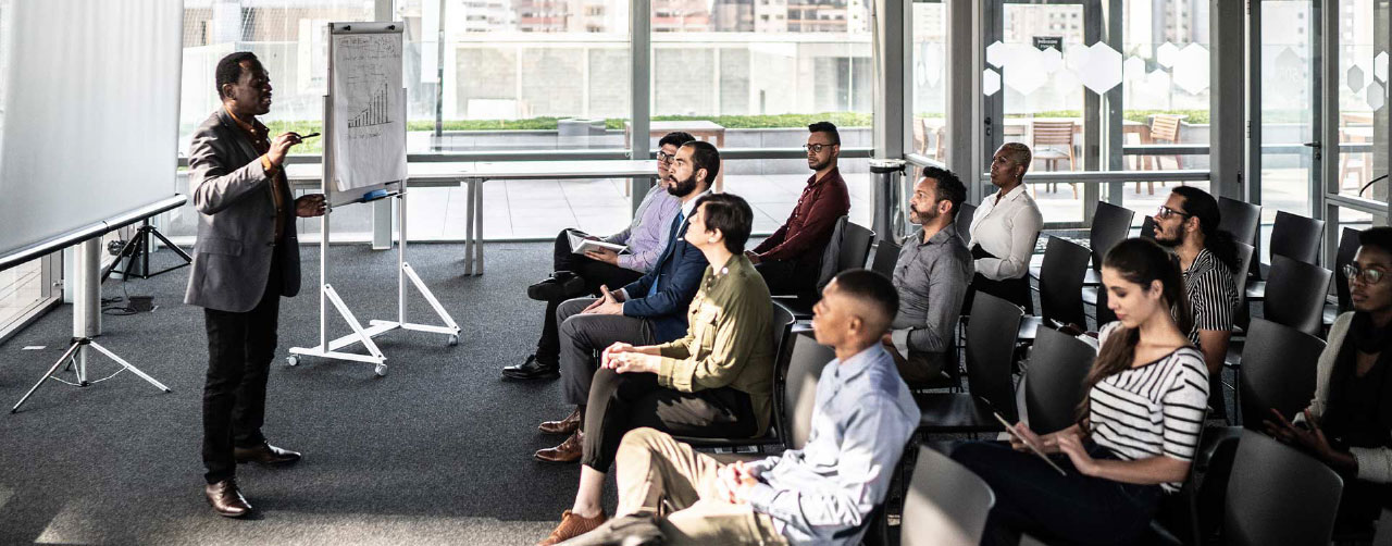 Modern bir sınıf veya seminer alanında, sunum yapan bir eğitmeni dinleyen yetişkin öğrenci grubunun yer aldığı eğitim ortamı.