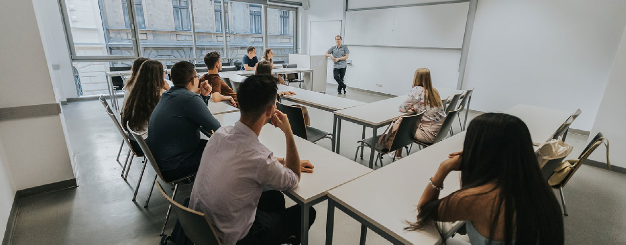 Aydınlık bir sınıfta, ders anlatan bir eğitmeni dinleyen öğrencilerin yer aldığı üniversite ders ortamı.